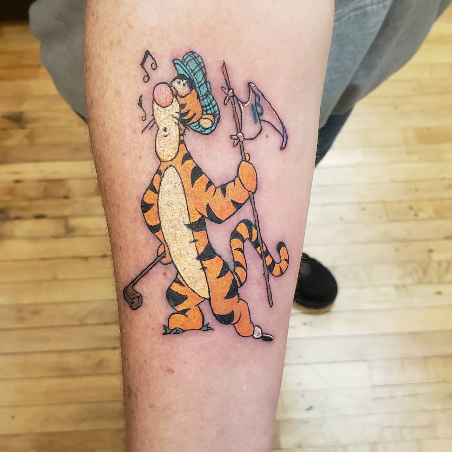Forearm tattoo of Tigger ice fishing, wearing a hat and holding a fishing rod with a fish